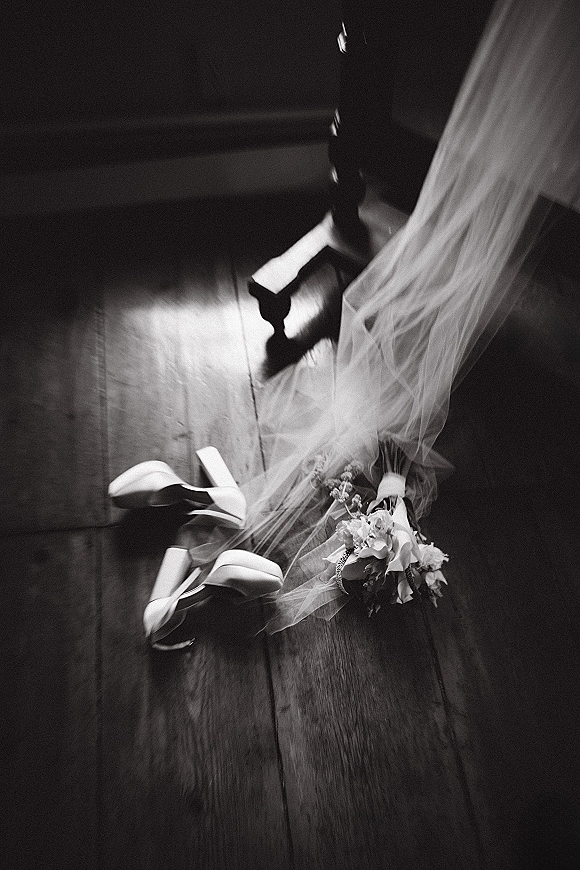 Bridal shoes in a wedding shoes flat lay with ankle strap heels, veil tulle and corsage on a wood floor in soft window light
