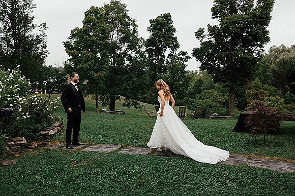 First look moment outdoors as bride in a strapless long-train gown walks up the stone path toward groom in a black tuxedo on a garden lawn