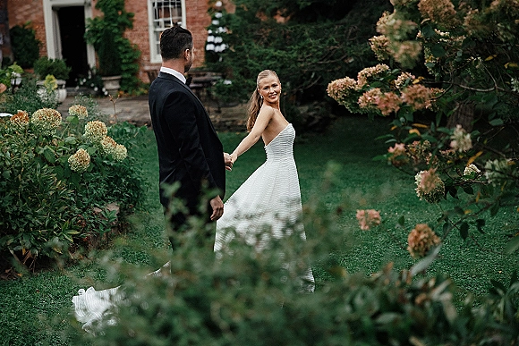 Couple portrait of bride and groom holding hands, bride looking back in strapless gown, groom in tuxedo on garden lawn by brick house