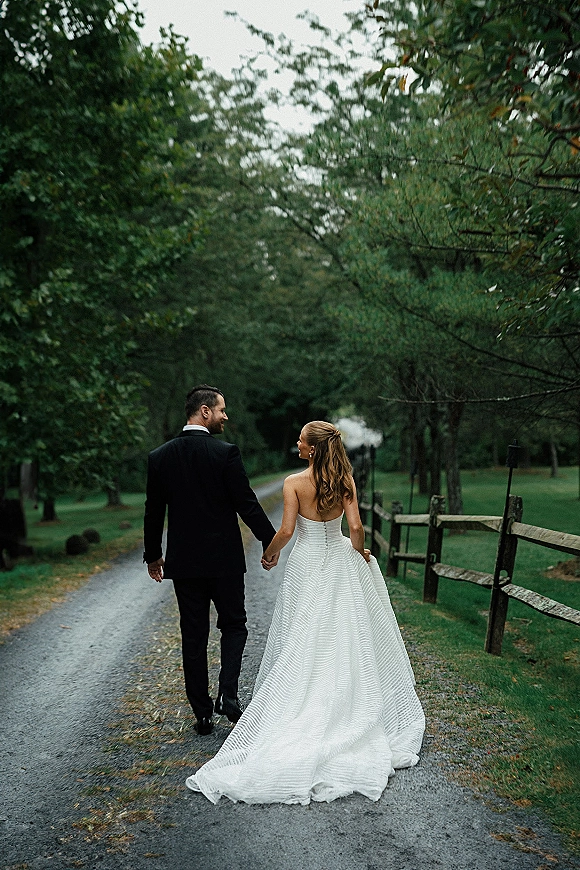 Couple portrait of newlyweds holding hands walking away, bride’s long train trailing on a tree-lined gravel path by a wooden fence