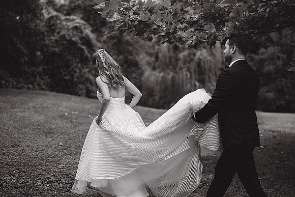 Wedding couple portrait of bride and groom walking away as he holds her gown train under a leafy garden canopy on the lawn