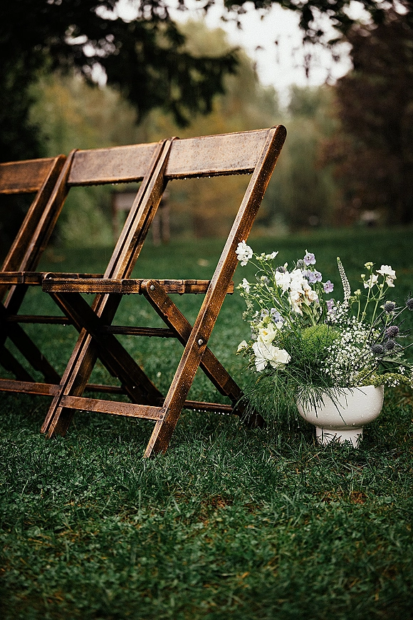 Ceremony chair decor with rustic wedding chairs and a pedestal vase of white and lavender flowers with greenery on a garden lawn
