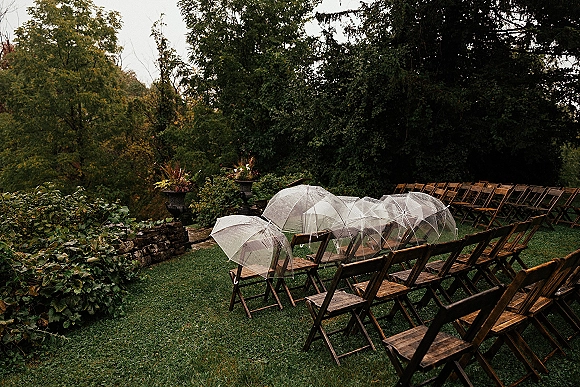 Outdoor ceremony seating with wood folding chairs and clear umbrellas lined on a lawn by a stone wall, trees, and garden foliage under gray sky