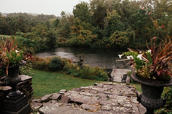 Wedding venue landscape at a lakeside wedding venue with stone steps, flower planters, and a wooden dock beside lawn and trees under overcast sky