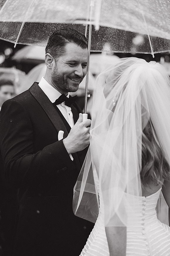 Wedding couple portrait of bride and groom under umbrella, her veil over her face, smiling in rain with blurred outdoor guests behind