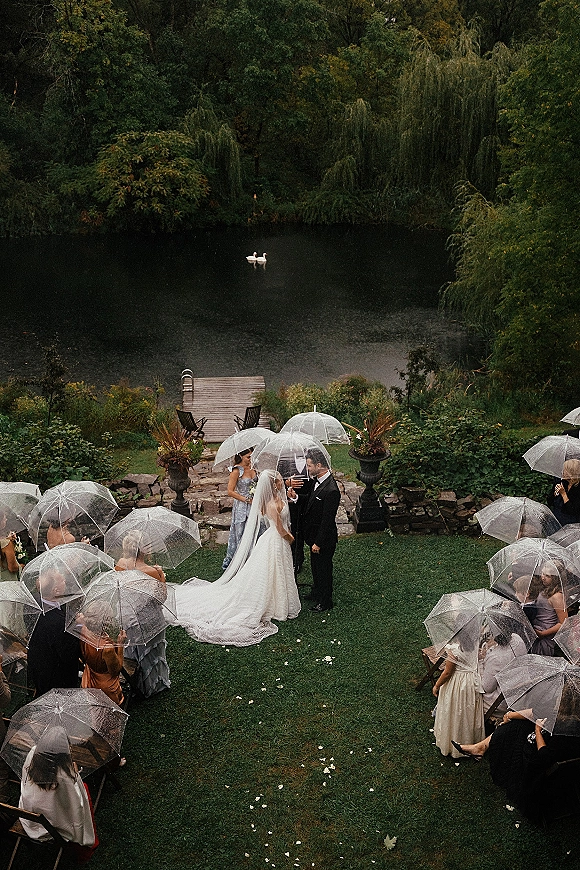 Outdoor wedding ceremony in the rain with clear umbrellas as the bride and groom stand on stone steps by a lakeside dock and lawn chairs