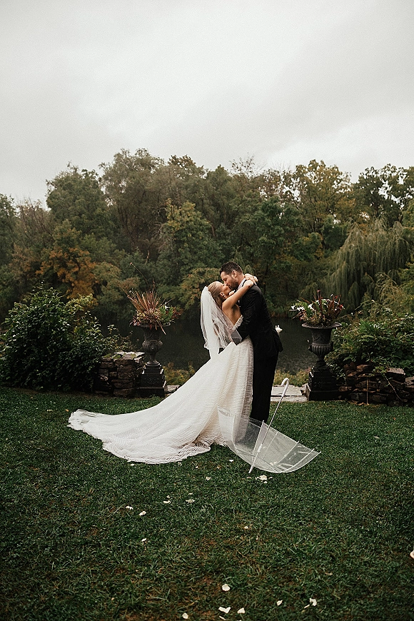 Wedding kiss portrait of bride and groom kiss under a clear umbrella, veil and long train flowing on a lakeside lawn by a stone wall