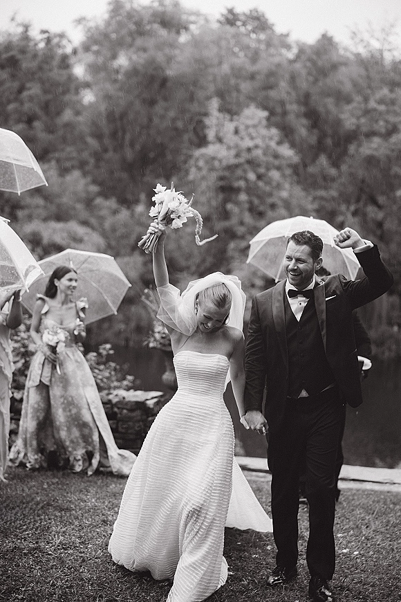 Wedding recessional as bride and groom walking hand in hand in rain, bride lifts bouquet, groom fist pumps under clear umbrellas outdoors