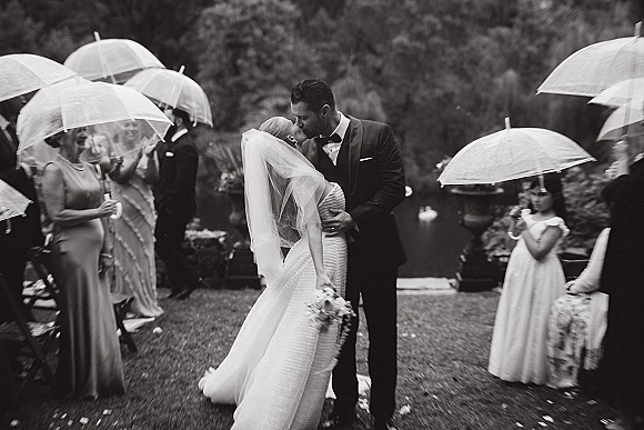 Wedding kiss in a rainy wedding kiss moment as bride and groom embrace under clear umbrellas, her veil and bouquet in a garden by water feature