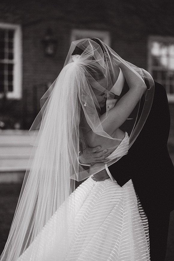 Wedding kiss portrait with veil over face kiss as bride and groom embrace under tulle veil, pearl earrings, brick facade behind