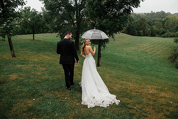 Couple portrait with a clear umbrella, bride in strapless gown with long train and groom in suit walking through grassy hills under overcast sky