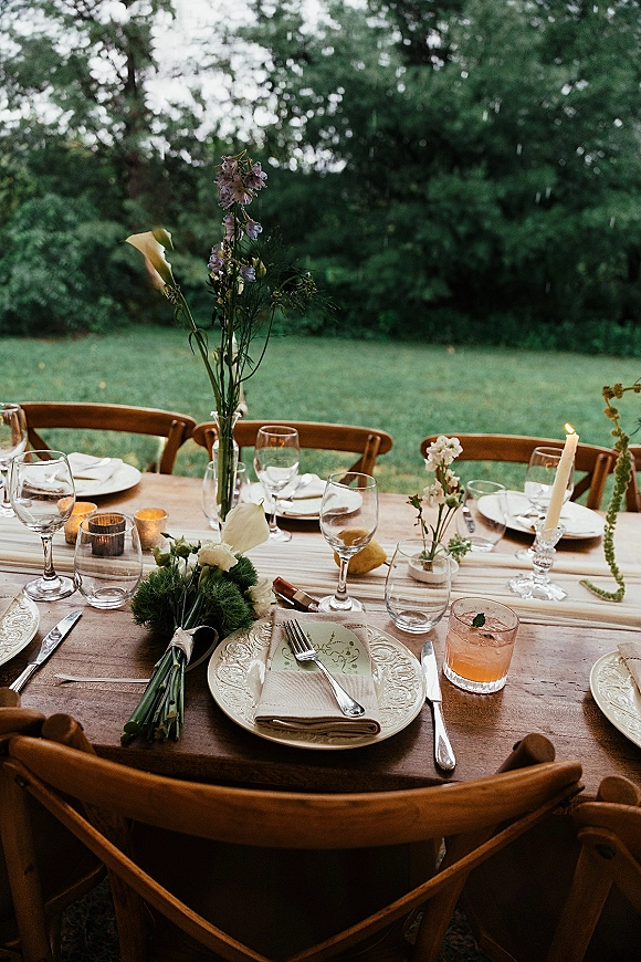 Reception tablescape with an outdoor reception table on a wood farm table, taper candles, bud vases, pears, and place cards on grass under trees