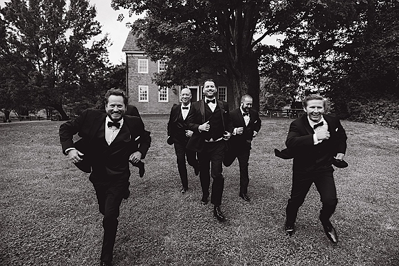 Groomsmen portrait of men running across a grass lawn in black tuxedos with bow ties, with large trees and a brick house behind them