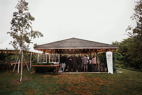 Cocktail hour setup with wedding outdoor bar, menu board and wood counter under a tent canopy with string lights on a grassy lawn