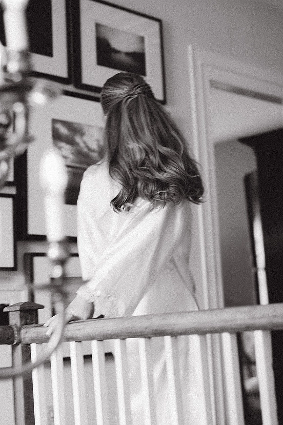 Bridal getting ready moment as bride in robe walks up a staircase, seen from behind with half-up curled hair by framed wall photos