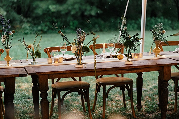 Reception tablescape with a farmhouse wedding table, wildflower bud vases, taper candles, and wine glasses under a tent on the lawn