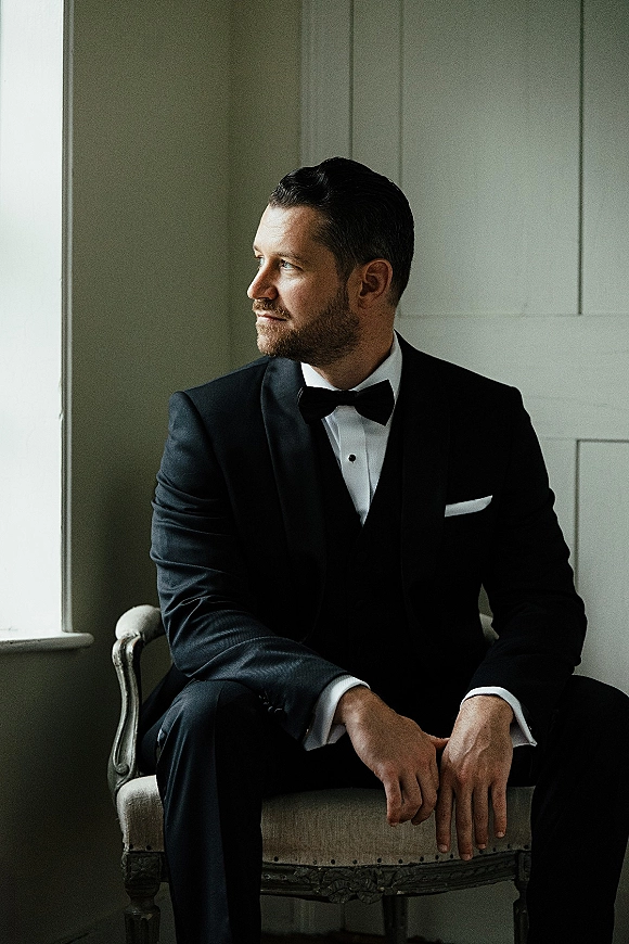 Groom portrait in a black tuxedo and bow tie, seated in an upholstered chair with wedding ring visible, lit by window light by a paneled wall