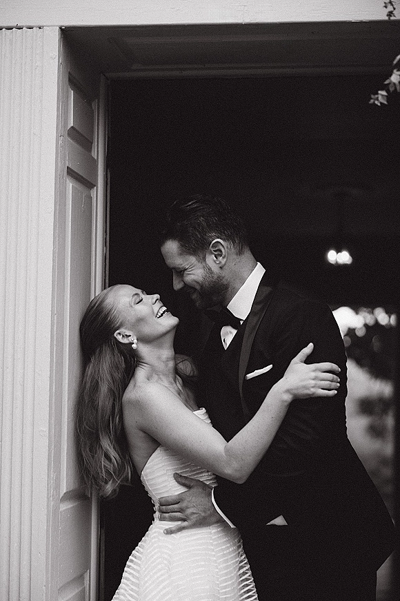 Couple portrait of bride and groom embracing and laughing in a doorway, her strapless dress and pearl earrings against bokeh lights