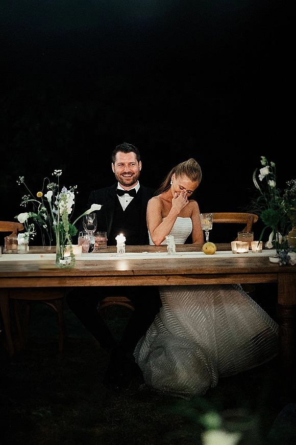 Wedding reception couple laughing at a candlelit sweetheart table, bride in strapless dress with groom in black tuxedo, dark greenery backdrop