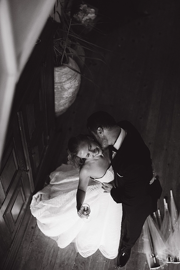 Wedding couple portrait in a black and white wedding photo, groom kissing bride’s cheek as she holds a champagne coupe in window light