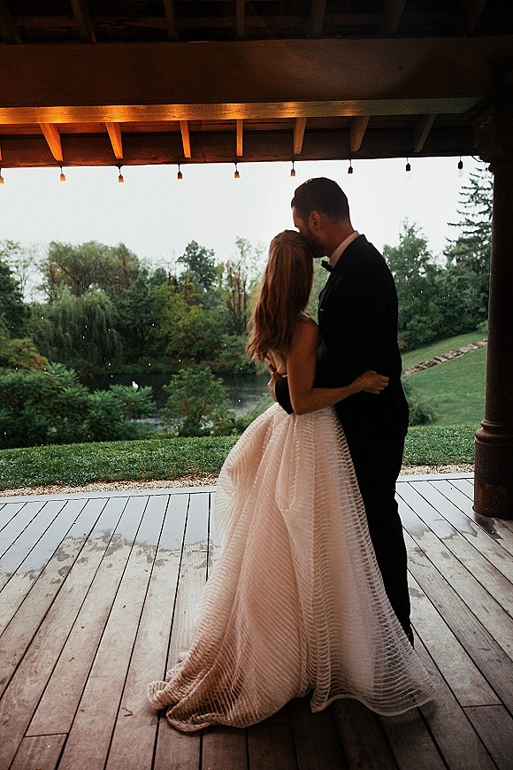 Wedding couple portrait of bride and groom embrace under string lights on a covered porch with trees, lawn, and pond beyond