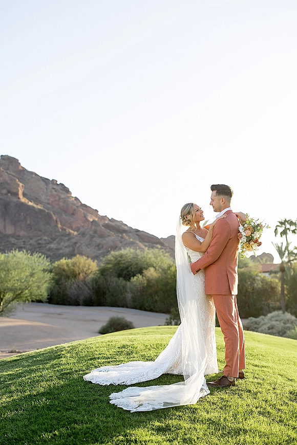 Couple portrait of bride and groom embracing outdoors, her veil and bouquet flowing, with mountains and desert landscape behind them