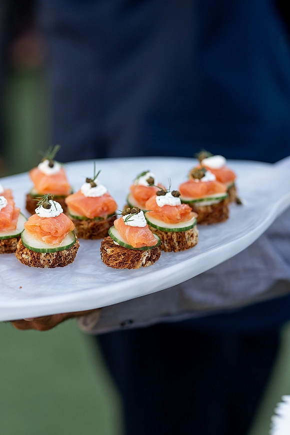 Wedding cocktail appetizers of smoked salmon canapes on crostini with cream cheese, cucumber, capers, and dill on a platter held by a suited caterer outdoors
