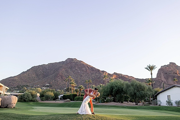 Wedding kiss portrait of bride in a white lace dress and groom in a tan suit dipping for a kiss with mountains and palm trees behind them