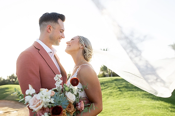 Couple portrait of bride and groom close up, smiling as she holds a colorful bouquet with veil in warm backlit sunlight on a lawn