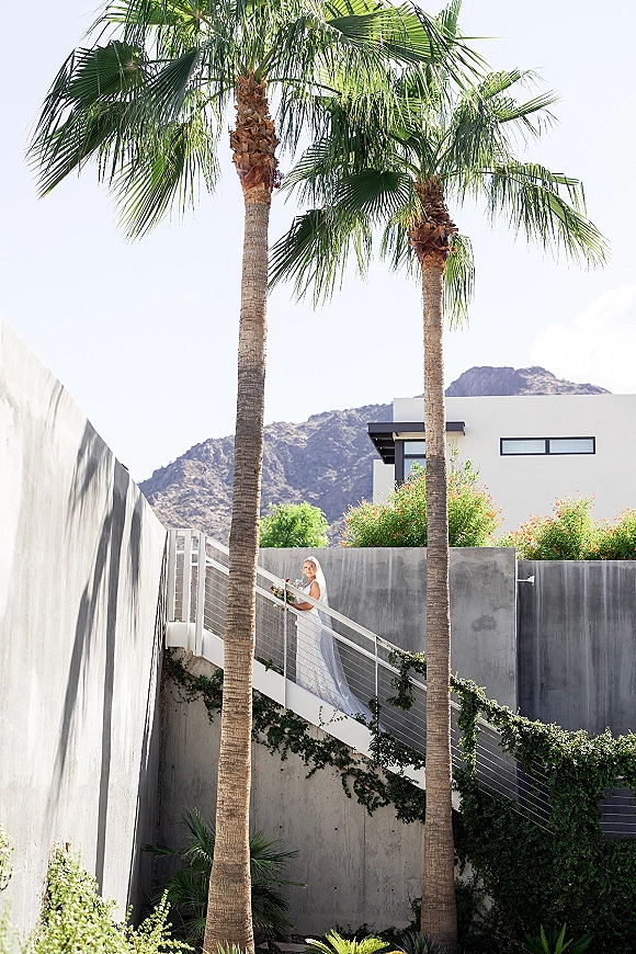 Bridal portrait of a bride holding bouquet, looking over her shoulder on an outdoor staircase with palm trees and mountain backdrop, veil trailing