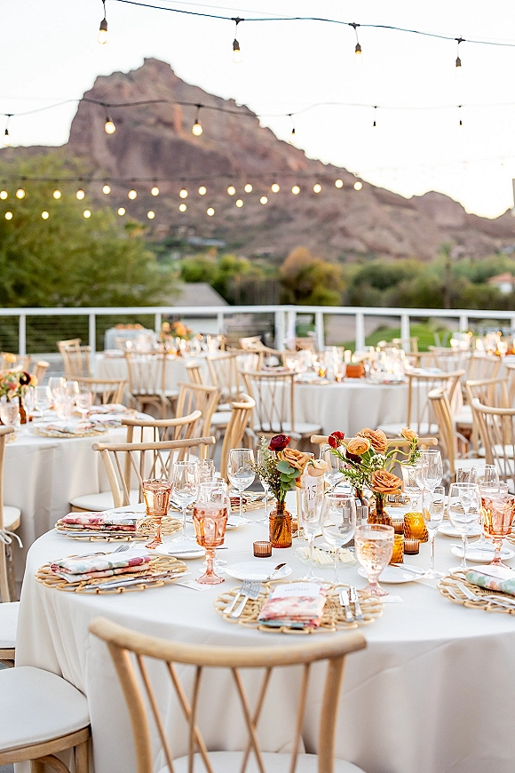 Reception tablescape at an outdoor wedding reception with white linens, amber bud vases, candlelight, and string lights framing mountain views