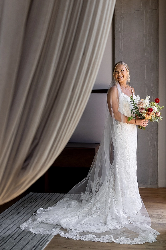 Bridal portrait of a bride holding bouquet in a lace gown with long veil, smiling over her shoulder in an indoor room with draped curtains
