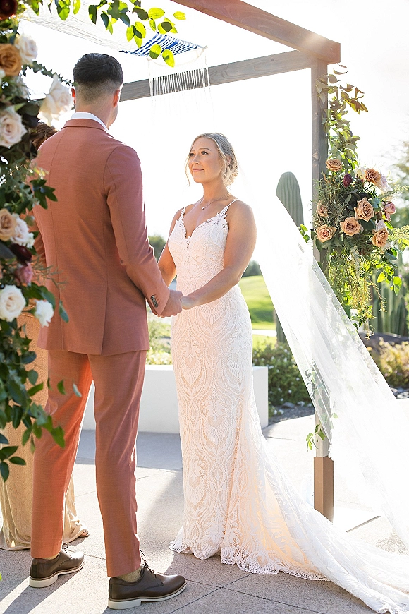 Wedding vows as bride and groom hold hands beneath a modern wooden wedding arch with rose greenery, sunlit lawn and trees behind