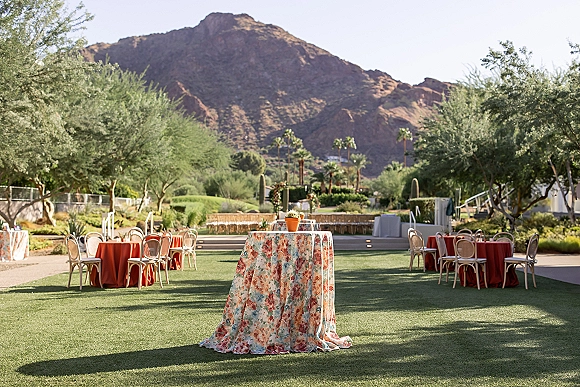 Outdoor reception setup with round tables and orange linens, bentwood chairs and potted centerpieces on a lawn with mountain backdrop