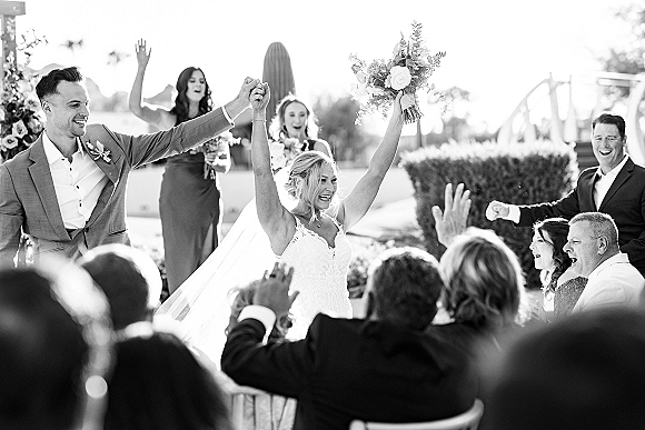 Recessional moment as bride and groom walk back down the aisle, bouquet raised, guests cheering beside garden ceremony chairs and hedges