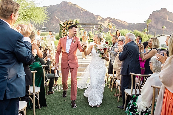 Wedding recessional with bride and groom walking aisle, holding hands as guests cheer and toss petals beneath a floral arch with mountains behind