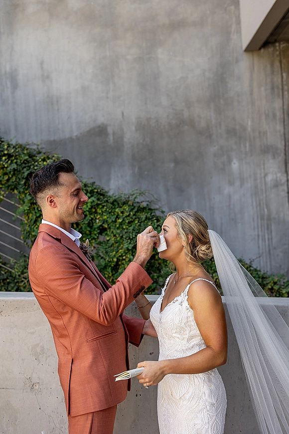 First look moment as groom wipes bride’s tears, her veil and lace dress visible, beside an ivy-lined concrete wall backdrop