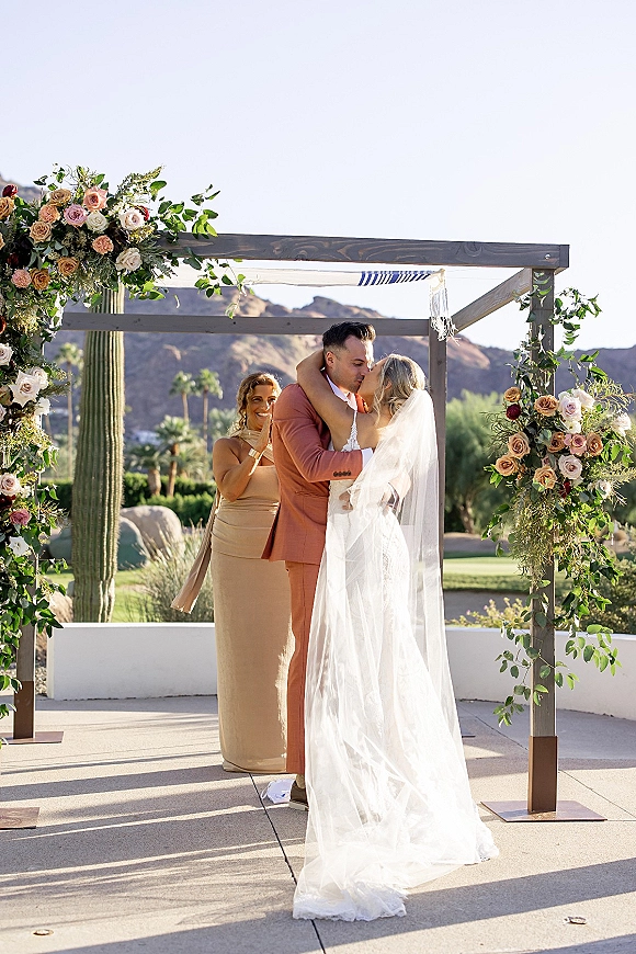 Wedding kiss beneath a floral arch, bride in lace gown and long veil with groom in suit, framed by desert mountains and cactus backdrop