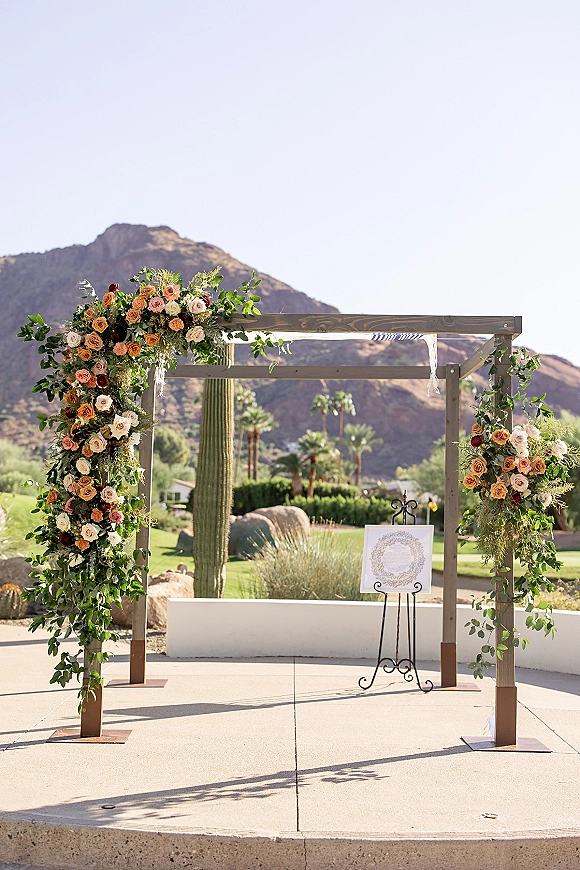 Wedding ceremony arch with roses and greenery on a wooden frame, set against desert mountains with cactus and palm trees under blue sky