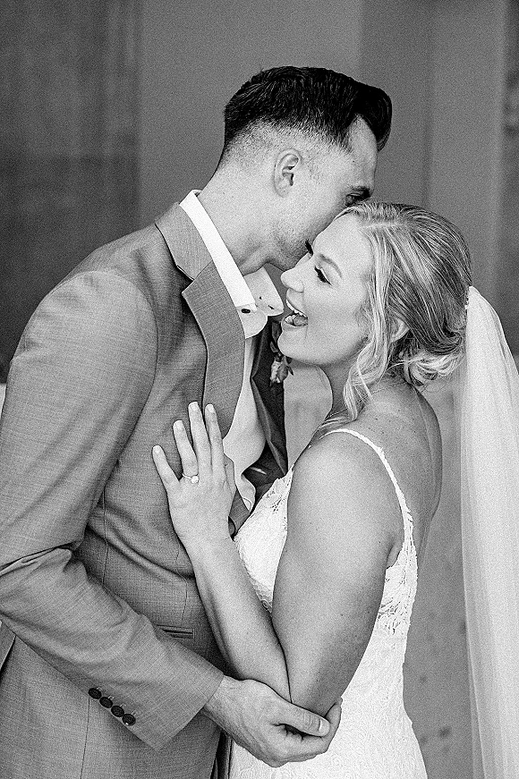 Wedding couple portrait of bride and groom embrace as he kisses her forehead, lace dress and veil visible against a stone wall backdrop