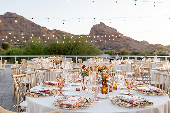 Reception tablescape with rattan charger plates, patterned napkins, floral centerpieces and amber votives on a deck overlooking mountains under string lights