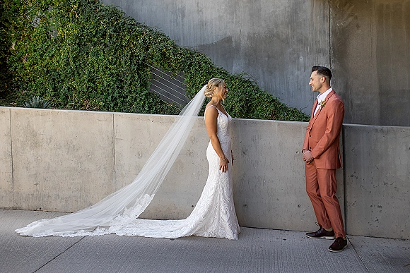 First look moment as bride in lace gown and long veil approaches groom in rust suit by a concrete wall with ivy slope backdrop