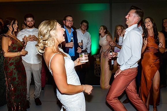 Wedding dance floor reception dance party with bride in lace dress and groom shirt laughing as guests cheer under green uplighting