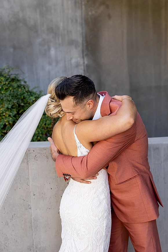 Wedding couple hug in a bride and groom embrace, her long veil over a lace dress as he holds her by a concrete wall with greenery