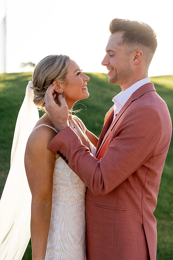 Couple portrait of bride and groom portrait as he adjusts her hair comb, lace dress and veil glowing in sunlight with green hills behind
