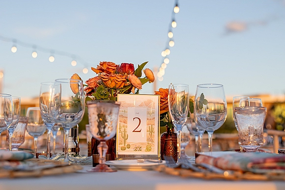 Reception tablescape with an outdoor wedding tablescape of orange rose centerpiece, candlelight, amber bottles, and string lights under open sky