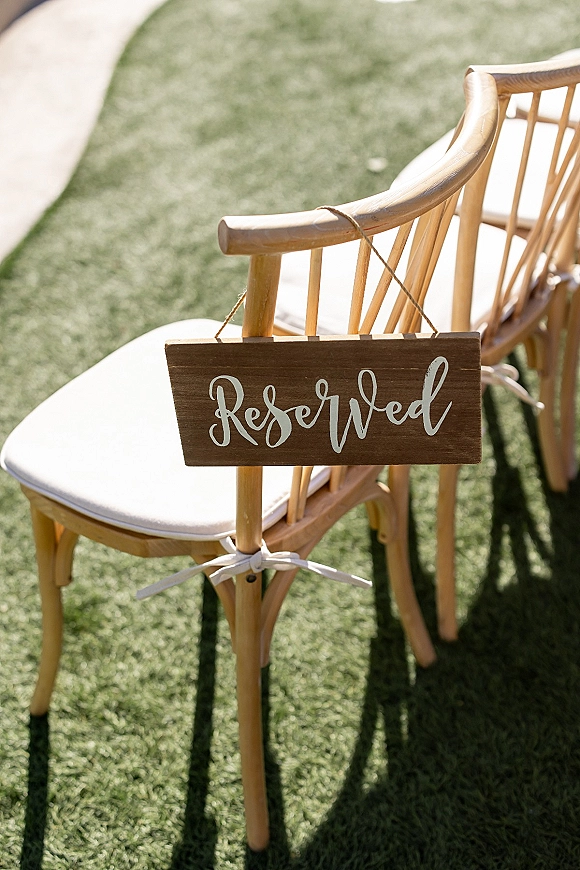 Wedding reserved sign with calligraphy lettering hanging by twine on a wooden chair, ribbon tied, on a sunlit lawn walkway