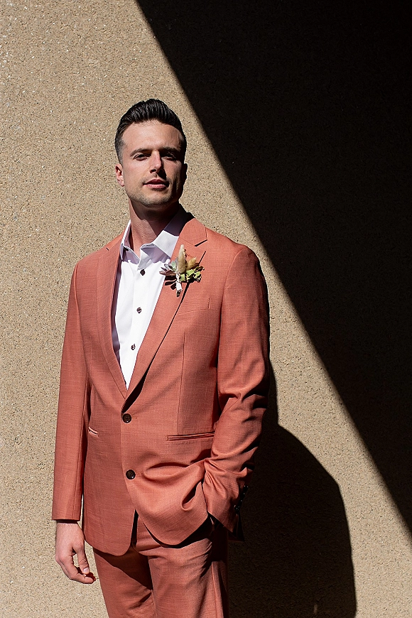 Groom portrait in a salmon wedding suit with white shirt and boutonniere, hands in pockets against a sunlit stucco wall with shadows