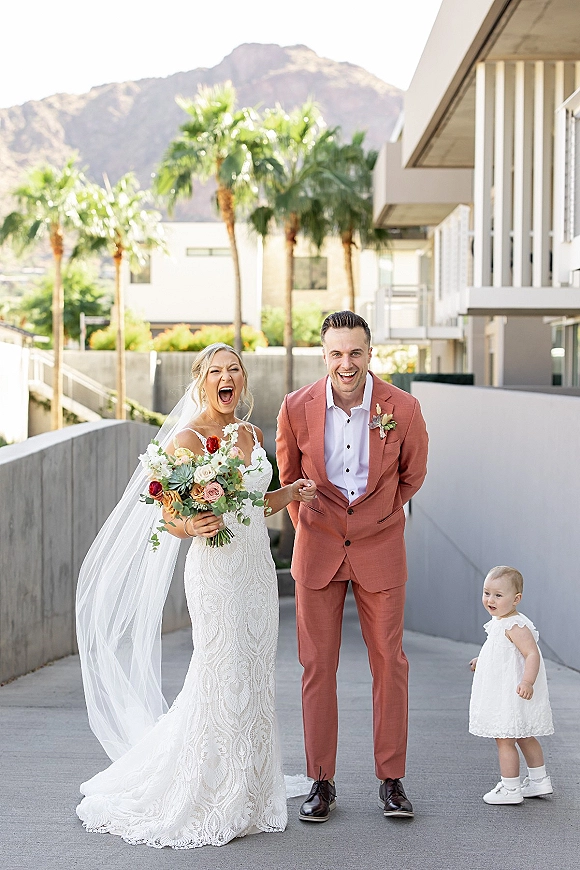 Wedding couple portrait of a laughing bride and groom holding hands, bride in lace dress with long veil, modern venue with palm trees and mountains behind