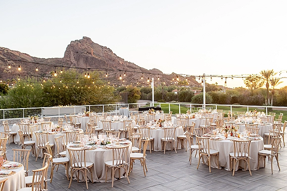 Outdoor wedding reception with reception tablescape of round tables, linen cloths and wooden chairs under string lights on a sunset patio with mountain views